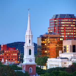 White church steeple with a clock tower in front of modern glass and brick office buildings at dusk, with a wooded hill behind